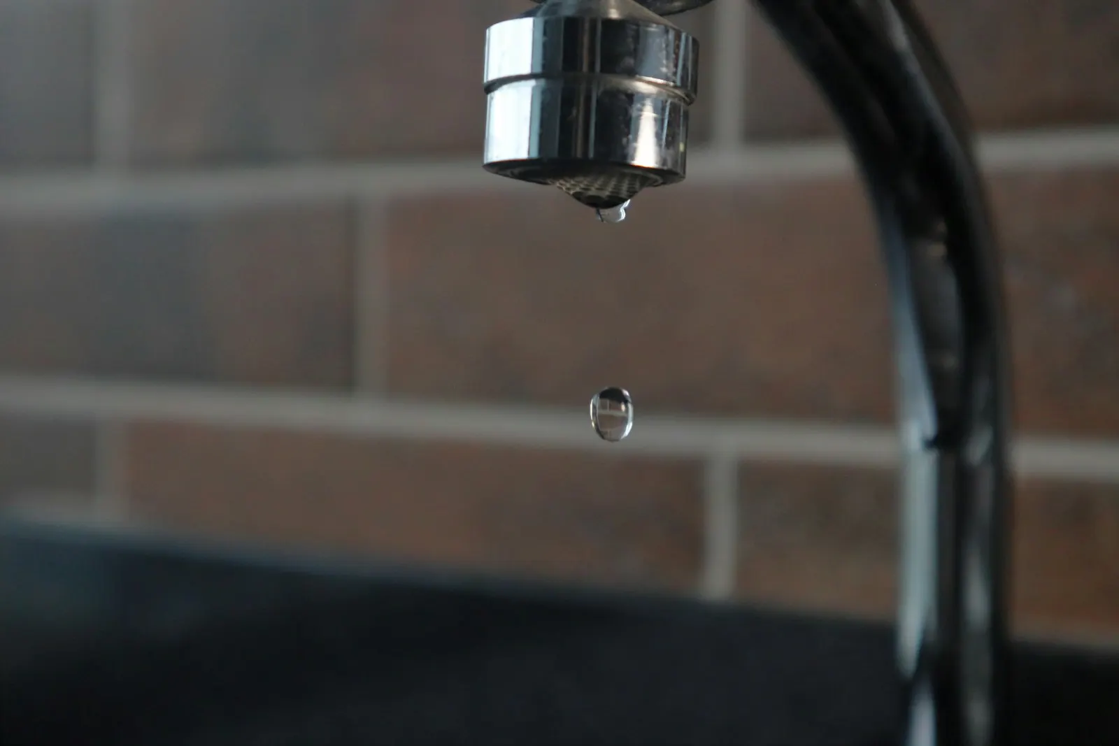 A drop of water falling from a chrome kitchen tap in front of a brick wall